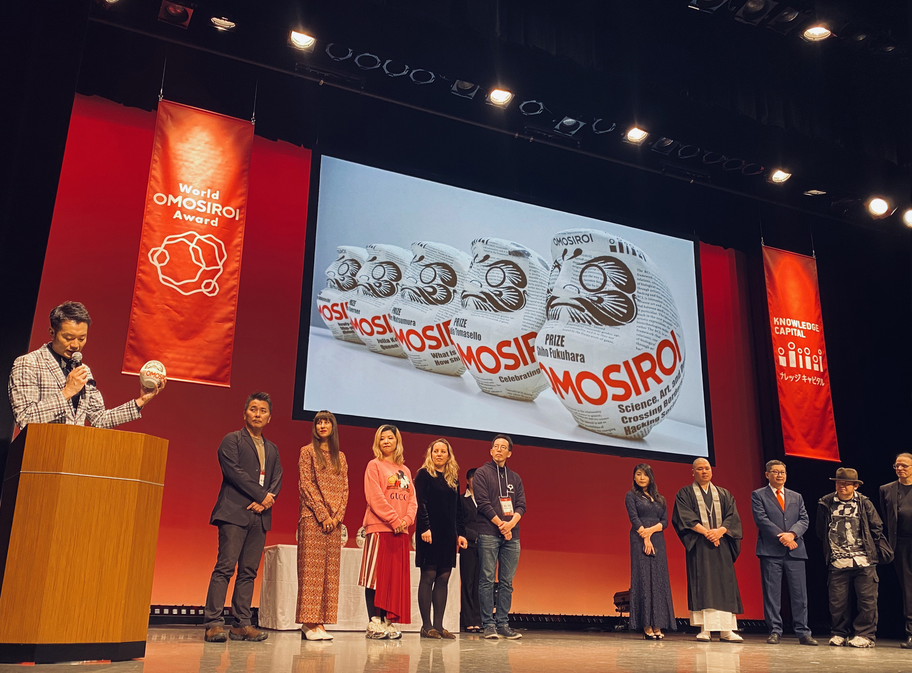 司会者がトロフィーのだるまアートを持っている写真