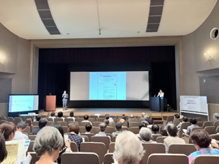 講演の様子。手話通訳者と講師が壇上にいる。手話通訳者の横には要約筆記のプロジェクターがある。